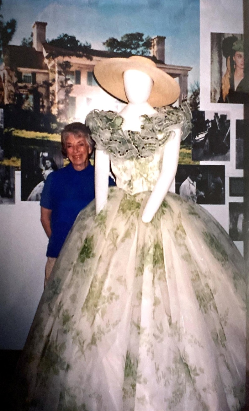 May include: A full-length gown featuring a white base with a green floral print, a voluminous skirt, and a ruffled neckline. The garment is displayed on a mannequin wearing a wide-brimmed straw hat. The dress has a fitted bodice and a full, layered skirt.