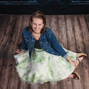 May include: A dark blue denim jacket worn over a black top and a white, knee-length, full-circle skirt featuring a green botanical leaf print. The outfit is paired with brown wedge sandals with ankle straps, creating a layered look.