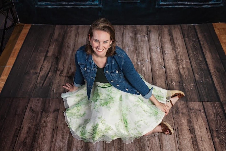 May include: A dark blue denim jacket worn over a black top and a white, knee-length, full-circle skirt featuring a green botanical leaf print. The outfit is paired with brown wedge sandals with ankle straps, creating a layered look.