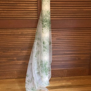 May include: A roll of sheer white fabric featuring a repeating botanical print in shades of green. The lightweight material is draped vertically, showcasing the delicate foliage pattern that covers the translucent surface of the textile roll.