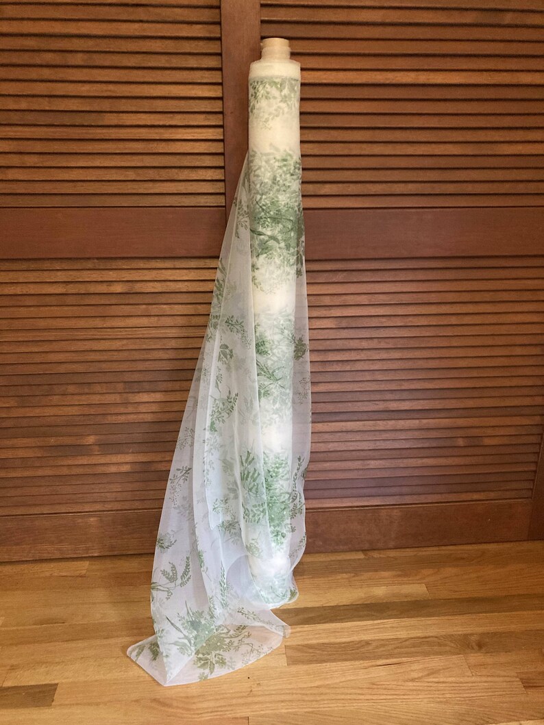 May include: A roll of sheer white fabric featuring a repeating botanical print in shades of green. The lightweight material is draped vertically, showcasing the delicate foliage pattern that covers the translucent surface of the textile roll.