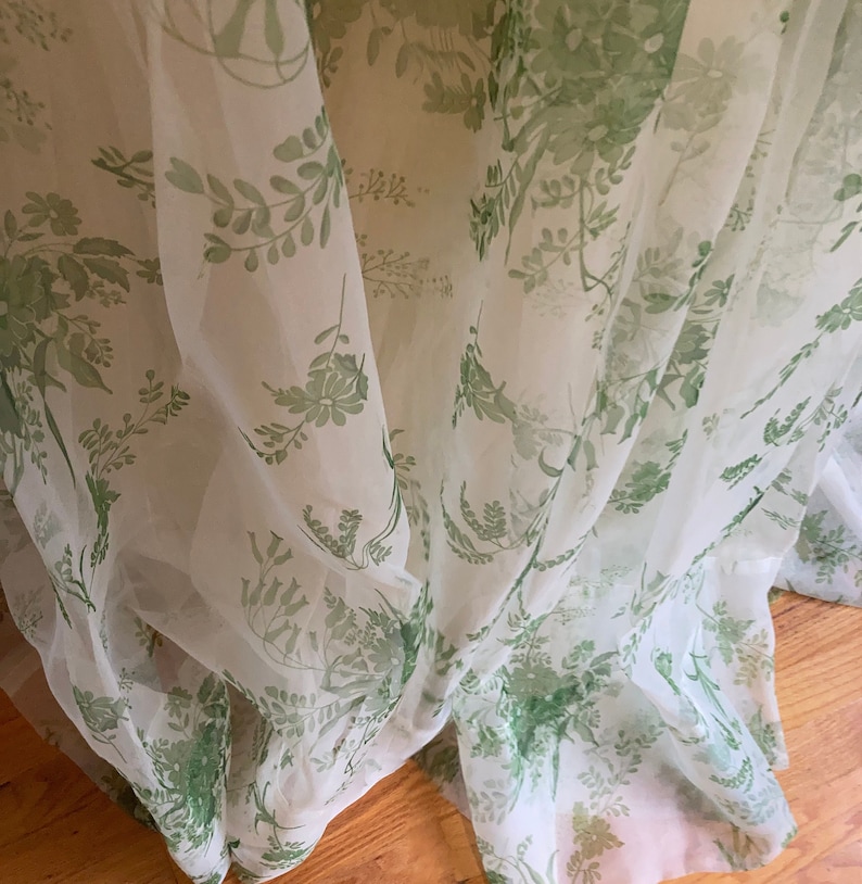 May include: A sheer, lightweight fabric garment featuring a delicate botanical print in shades of green on a white background. The material is draped in soft folds, showcasing a repeating pattern of various leaves, stems, and small flowers.