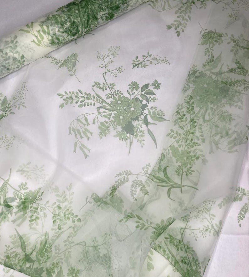 May include: A sheer white fabric roll featuring a repeating botanical print in shades of sage green. The pattern consists of delicate floral bouquets and trailing leafy vines scattered across the translucent material, creating a soft layered effect.