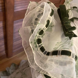 May include: A sheer white organza garment featuring a green floral print, layered ruffled edges, and dark green velvet ribbon trim. A small dark green velvet bow is attached to the upper section, accentuating the layered, translucent fabric texture.