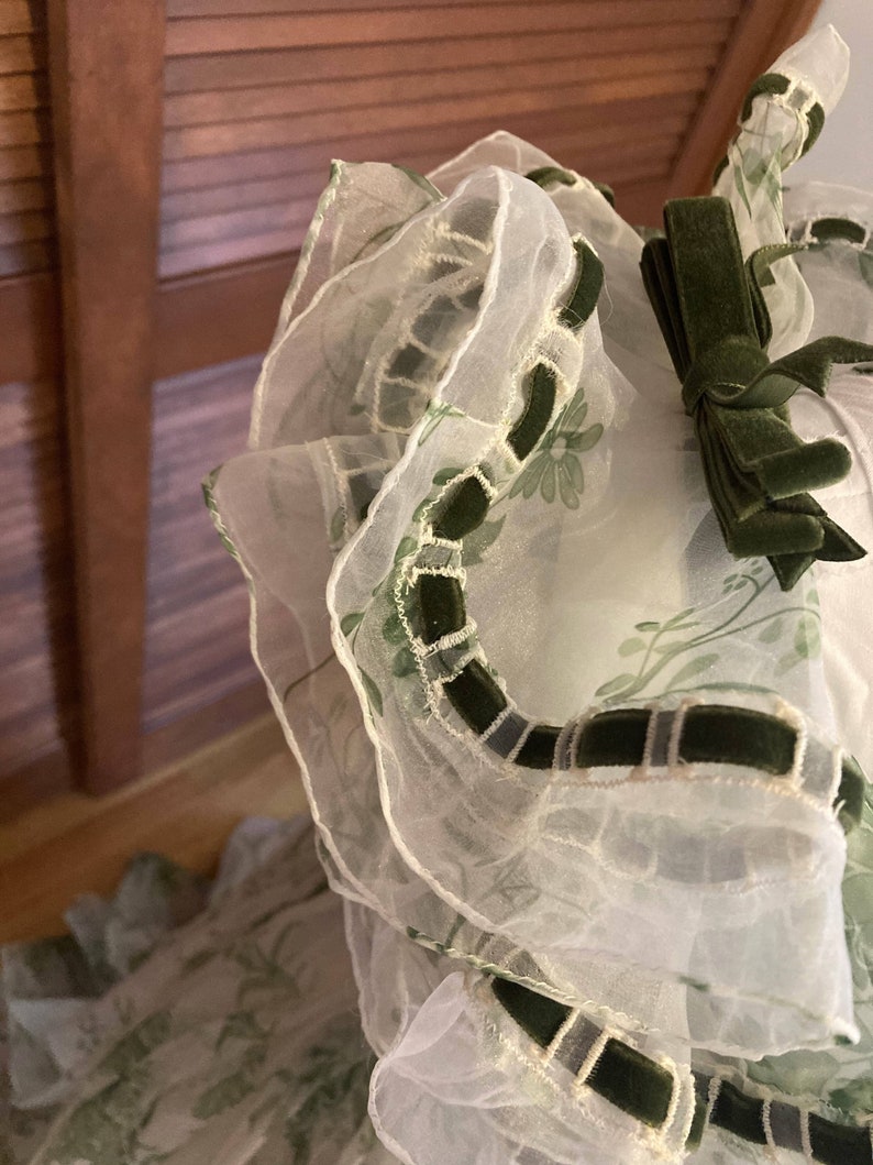 May include: A sheer white organza garment featuring a green floral print, layered ruffled edges, and dark green velvet ribbon trim. A small dark green velvet bow is attached to the upper section, accentuating the layered, translucent fabric texture.
