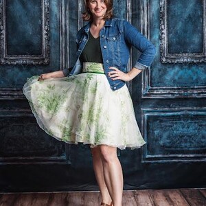 May include: A blue denim jacket worn over a dark green v-neck top and a white knee-length skirt featuring a green botanical print. The skirt has a wide waistband and a flared silhouette. The outfit is paired with brown wedge sandals with straps.