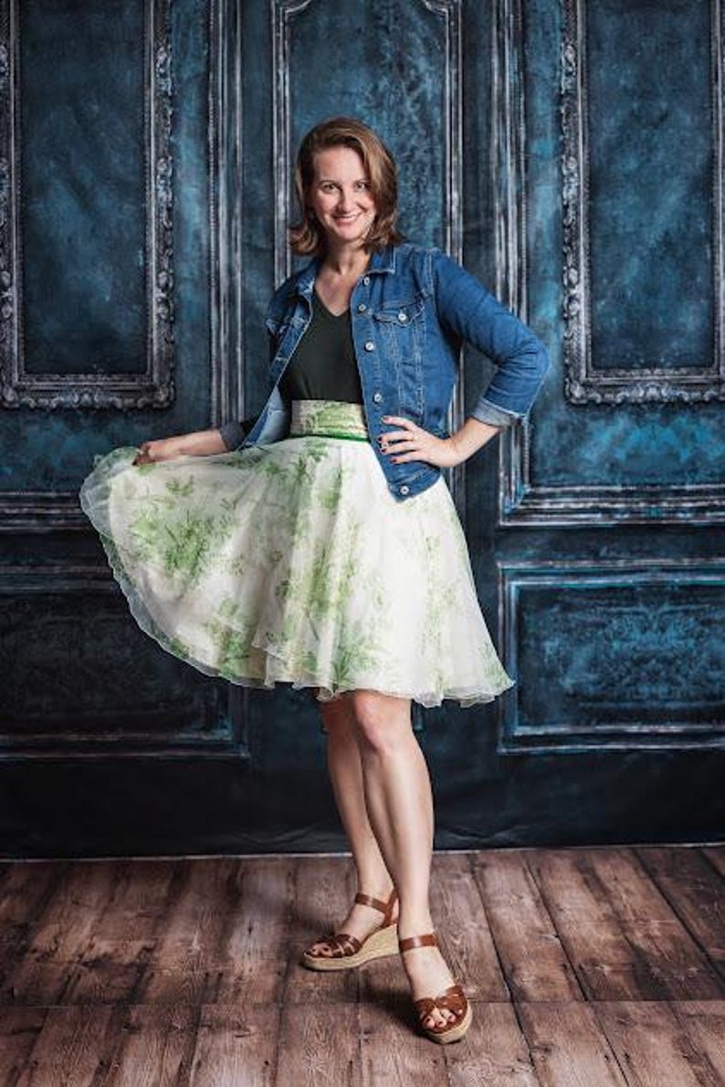 May include: A blue denim jacket worn over a dark green v-neck top and a white knee-length skirt featuring a green botanical print. The skirt has a wide waistband and a flared silhouette. The outfit is paired with brown wedge sandals with straps.