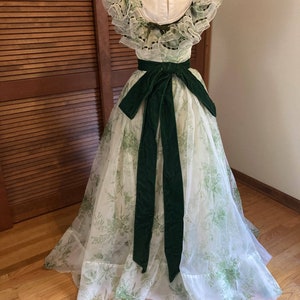 May include: A floor-length dress featuring a white sheer overlay with a green botanical print, ruffled cap sleeves, and a high neckline. A wide, dark green velvet sash is tied in a bow at the waist, with long tails extending down the full skirt.