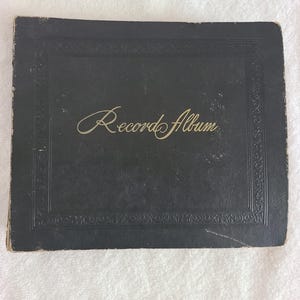 May include: A vintage black record album with the words "Record Album" in cursive script. The album has a decorative border and shows signs of wear, suggesting it is an antique or a well-loved item.