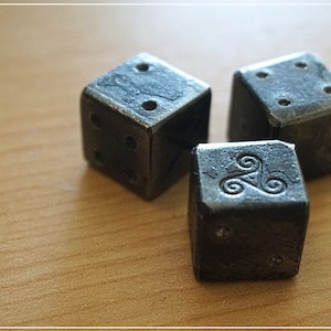 May include: Three black metal dice with different symbols on each side. The dice are on a light brown wooden surface.