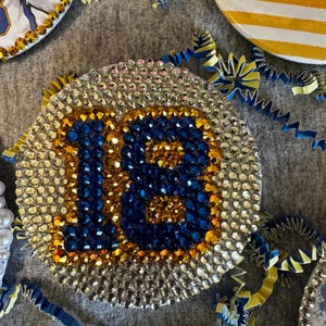 Op de afbeelding: Een ronde decoratieve plaquette met het getal 18 prominent in het midden weergegeven. Het getal is gemaakt van donkerblauwe en gouden strass-steentjes, terwijl de achtergrond is gevuld met heldere, glinsterende strass-steentjes.