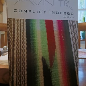 May include: A book cover with the title "CONFLICT INDEEGO" by Kblaze. The cover features a colorful woven pattern in shades of green, red, white, and black. Above the title is a stylized mountain range graphic.