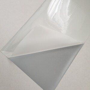 May include: A sheet of clear adhesive vinyl with a grey adhesive backing. The vinyl is designed for use in crafting and other projects.