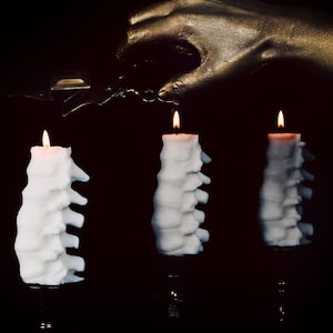 Pu&ograve; includere: Tre candele bianche con un design unico, testurizzato, a forma di spina dorsale, sono accese su uno sfondo nero. Una mano dipinta d'oro tiene un accendino sopra le candele.