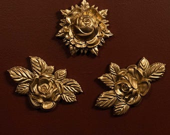 Puede incluir: Tres apliques florales metálicos de color dorado con rosas y hojas detalladas. La pieza superior es una rosa circular, mientras que las dos piezas inferiores son rosas acompañadas de conjuntos de hojas con textura.