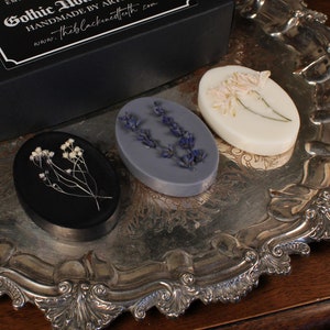 Könnte beinhalten: Ovale Seifen mit getrockneten Blumen und Kräutern auf einem silbernen Tablett, neben einer schwarzen Schachtel. Die Seifen sind schwarz, lavendelfarben und weiß, mit floralen Akzenten. Auf der Schachtel steht "Gothic Home Decor" und "Handmade by Artisans".