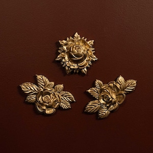 May include: Three gold-coloured rose ornaments with detailed petals and leaves are arranged on a dark brown background. One rose is centered, while the other two are positioned below, creating a decorative arrangement.