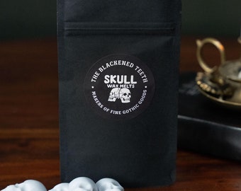 May include: A black pouch with a white sticker that says "The Blackened Teeth Skull Wax Melts Makers of Fine Gothic Goods". There are four white skull-shaped wax melts in front of the pouch.