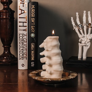 Pu&ograve; includere: Una candela bianca a forma di colonna vertebrale umana, che brucia su un vassoio di metallo dorato. La candela &egrave; davanti a una libreria con tre libri, uno intitolato "A Graveside Companion" e l'altro intitolato "Murder Isn't Easy".