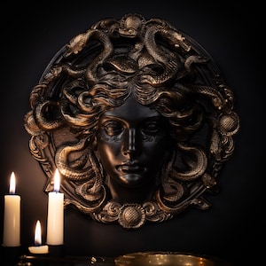 Könnte beinhalten: Schwarze und goldene Medusa-Wandskulptur mit detaillierten Schlangen und Gesichtszügen. Die Skulptur ist an einer dunklen Wand angebracht, mit drei brennenden Kerzen im Vordergrund.