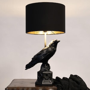 Può includere: Una lampada da tavolo con una base a forma di corvo nero poggiata su un piedistallo ottagonale. La lampada presenta un paralume cilindrico in tessuto nero con rivestimento interno dorato, sostenuto da un'asta centrale metallica sottile.