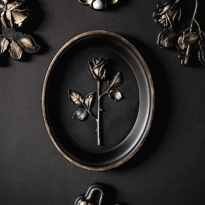 Puede incluir: Decoración mural ovalada negra con un diseño de rosa en relieve en el centro. La rosa y las hojas son doradas, sobre un fondo negro. El marco tiene un borde dorado. Se ven otras decoraciones con temática de rosas.