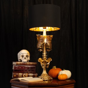 May include: A table lamp featuring a gold-colored human skeletal torso base, a black cylindrical lampshade with a gold interior, a skull with the text Memento Mori, a stack of four books, and three small pumpkins arranged on a wooden surface.