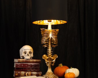 Könnte beinhalten: Eine Tischlampe mit einem goldfarbenen menschlichen Skelettorso als Basis, einem schwarzen zylindrischen Lampenschirm mit goldener Innenseite, einem Schädel mit der Aufschrift Memento Mori, vier Büchern und drei kleinen Kürbissen auf einer Holzfläche.