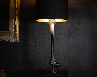 May include: A table lamp featuring a black cylindrical lampshade and a base shaped like a human femur bone. The bone structure transitions from a dark black at the bottom to a translucent, light-colored top, mounted on a square black base.