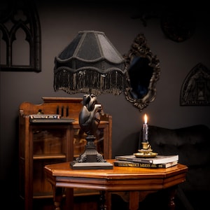 Puede incluir: Una lámpara de mesa con una pantalla de tela negra con flecos decorativos y una base esculpida con la forma de un corazón humano anatómico. La lámpara tiene una base cuadrada y un acabado en color negro mate uniforme en toda su estructura.