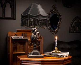 May include: A table lamp featuring a black fabric lampshade with decorative fringe and a base sculpted in the shape of an anatomical human heart. The lamp has a square pedestal base and is finished in a solid matte black color throughout.