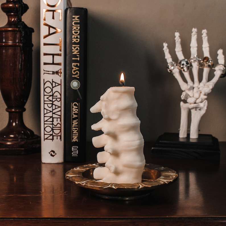 Pu&ograve; includere: Una candela bianca a forma di colonna vertebrale umana, che brucia su un vassoio di metallo dorato. La candela &egrave; davanti a una libreria con tre libri, uno intitolato "A Graveside Companion" e l'altro intitolato "Murder Isn't Easy".