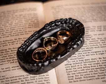 Puede incluir: Un cuenco ovalado de color negro con un borde texturizado que recuerda a una dentadura. Dentro del recipiente, tres anillos de metal dorado con diseños variados, incluyendo una banda lisa y dos piezas decoradas, descansan en el fondo.