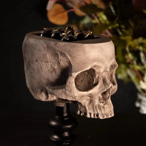 May include: A white ceramic skull ring holder with a black base. The skull is detailed with realistic features and has a hollow top to hold rings.