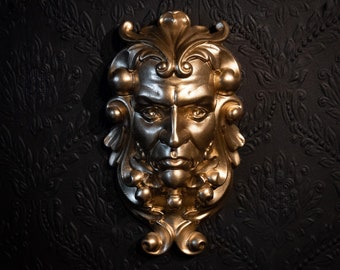 May include: A gold-colored decorative wall plaque featuring a stylized human face with intricate details. The face has a serious expression and is surrounded by ornate floral designs.