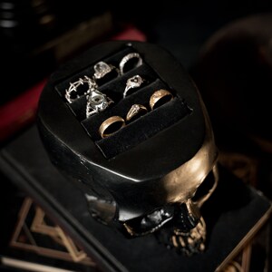 May include: A black skull-shaped ring holder with gold accents. The holder has five slots for rings, and is filled with various rings, including gold, silver, and gemstone rings.