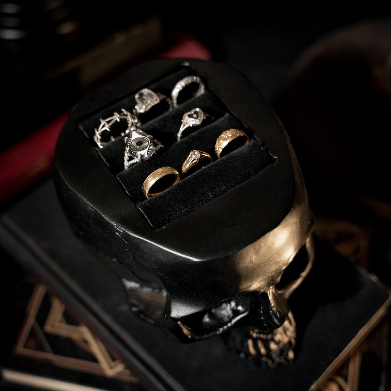 May include: A black skull-shaped ring holder with gold accents. The holder has five slots for rings, and is filled with various rings, including gold, silver, and gemstone rings.