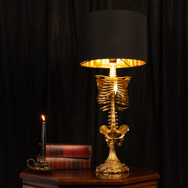 Puede incluir: Una l&aacute;mpara de mesa con una caja tor&aacute;cica y columna vertebral humana de color dorado como soporte central, rematada con una pantalla cil&iacute;ndrica negra que tiene un forro interior de color dorado met&aacute;lico. La base es ornamentada y plana.