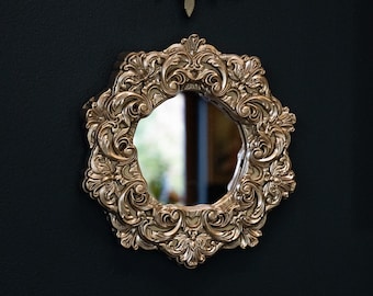 May include: A gold-toned ornate mirror with a round, decorative frame. The mirror reflects a blurry image of a room.