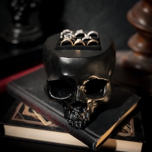 May include: A black skull ring holder with a gold finish. The skull has a detailed design and is sitting on top of two books.