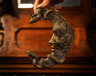 May include: A crescent moon-shaped wall hanging with a metallic bronze finish, featuring an embossed human face profile and two snakes coiled around the tips. The surface displays intricate, fine-line engravings and textured details throughout.