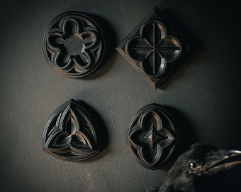 May include: Four dark, gothic-style wall decorations with intricate patterns are displayed against a dark gray wall. The shapes include a circle, a diamond, a triangle, and an oval. A black crow is in the foreground.