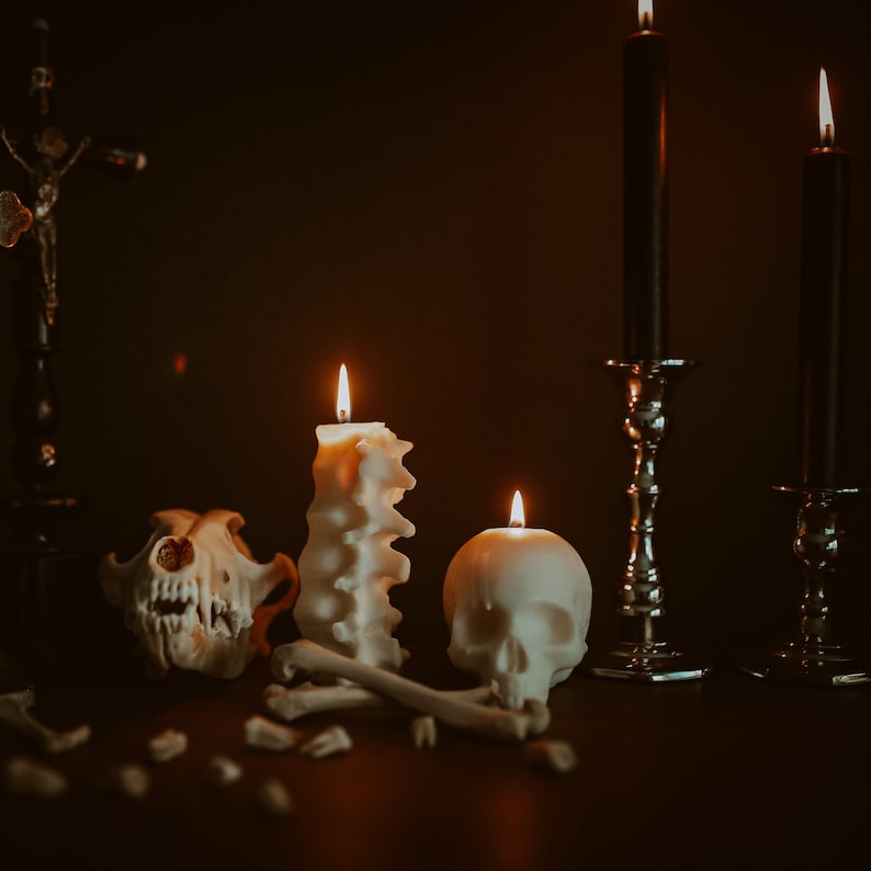 Pu&ograve; includere: Tre candele bianche, una a forma di colonna vertebrale, un'altra a forma di teschio e una terza a forma di candela tradizionale, sono accese in una stanza buia. Le candele sono su un tavolo con un teschio di animale bianco e ossa sparse intorno.