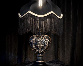 May include: A black table lamp with a decorative base and a fringed lampshade. The base is ornate with gold accents, and the shade is trimmed with white and black fringe. The lamp is a vintage-inspired design.