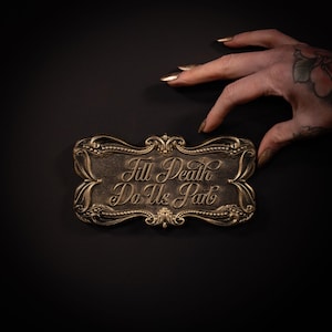 May include: A rectangular plaque with ornate, raised gold-toned borders and a dark, textured center. The text 'Till Death Do Us Part' is embossed in a flowing, elegant script font across the middle of the plaque, creating a high-contrast look.