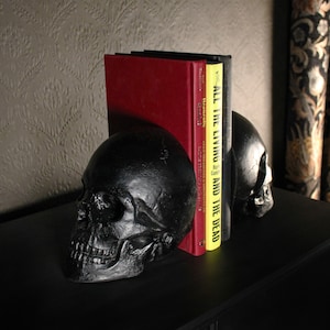 May include: Two black skull bookends with a red and yellow book titled "All the Living and the Dead" by Hay Cain.