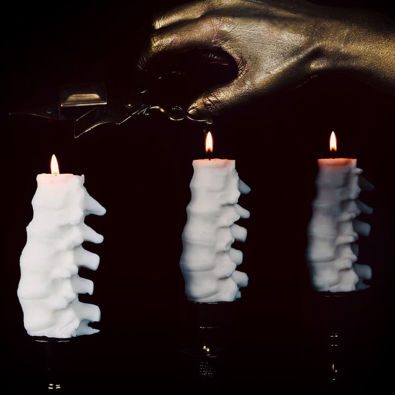 Pu&ograve; includere: Tre candele bianche con un design unico, testurizzato, a forma di spina dorsale, sono accese su uno sfondo nero. Una mano dipinta d'oro tiene un accendino sopra le candele.