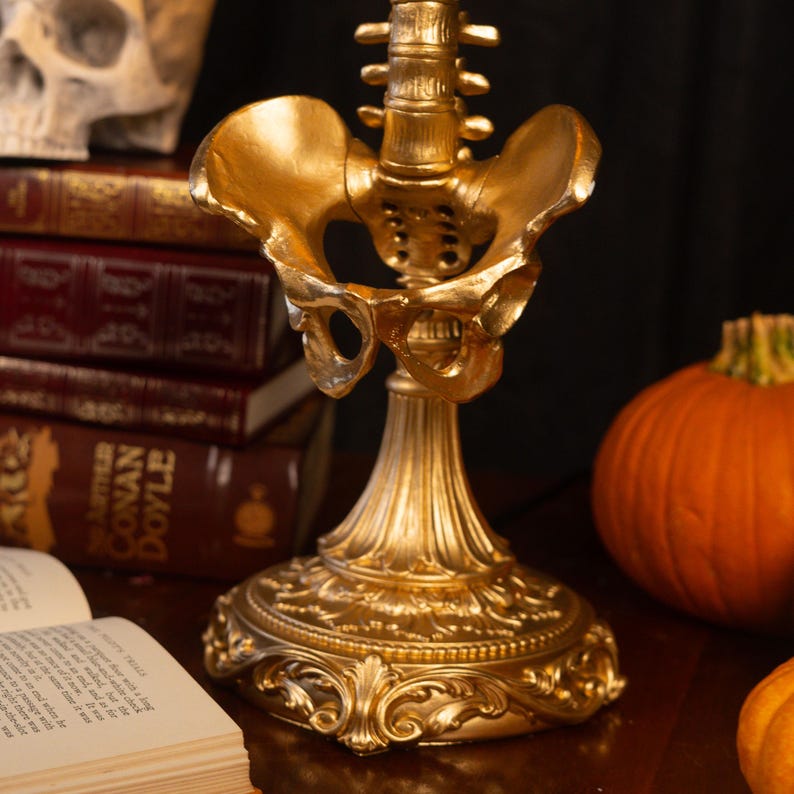 Puede incluir: Una escultura decorativa de color dorado que presenta una pelvis humana y una parte de la columna lumbar, montada sobre una base ornamentada estilo pedestal con detalles de volutas intrincados, acabada con un brillo met&aacute;lico en toda la estructura.