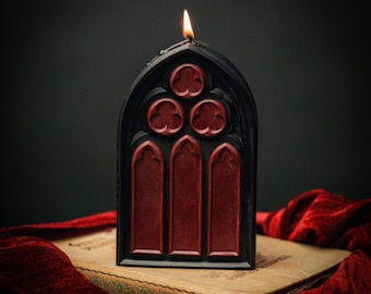 Könnte beinhalten: Eine schwarze und rote Kerze in Form eines gotischen Kathedralenfensters. Die Kerze ist angezündet und die Flamme ist sichtbar.