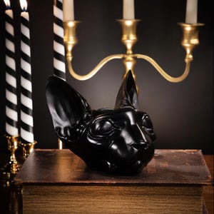May include: A glossy black cat head sculpture, resting on a vintage book. The sculpture is a detailed representation of a Sphynx cat. Two black and white striped candles and a gold candelabra with lit candles are in the background.